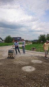 Drei Männer, darunter ein Schüler der AKS, arbeiten auf einer Baustelle, einer bedient einen Bagger, ein anderer schaufelt Erde, und der dritte beobachtet.
