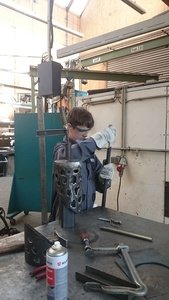Ein Schüler in blauer Jacke und weißen Handschuhen arbeitet in einer Werkstatt, umgeben von Werkzeugen und Geräten, an einer Metallskulptur.